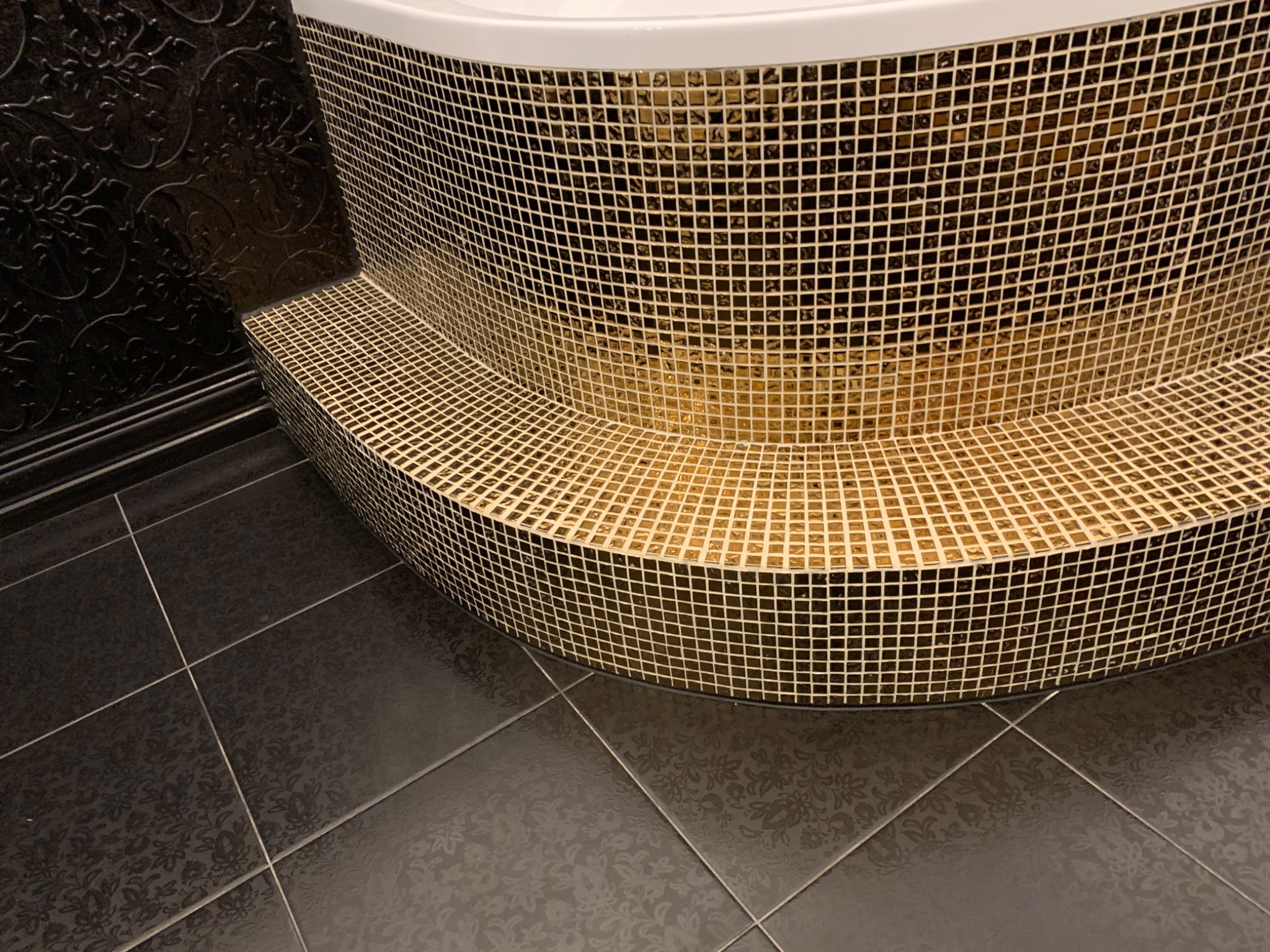 Mozaika w odcieniach złota i czerni zdobi obudowę wanny, kontrastując z ciemną, wzorzystą podłogą i ścianą, tworząc elegancki efekt wizualny.