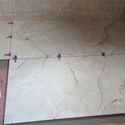 Usługi budowlane Vladysław Kliatskyi - Proces układania beżowych płytek podłogowych imitujących marmur w łazience, z użyciem poziomicy, przyssawek do przenoszenia płytek i systemu poziomowania z czerwono-niebieskimi klipsami.