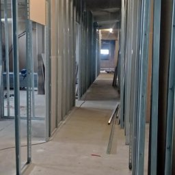 Usługi budowlane Vladysław Kliatskyi - Korytarz w budynku w trakcie remontu, widoczne metalowe profile konstrukcyjne ścian działowych z częściowo zamontowanymi płytami. Świecąca lampa sufitowa w tle.