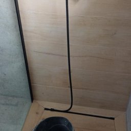 Usługi budowlane Vladysław Kliatskyi - Fragment łazienki w trakcie remontu: widoczny czarny zestaw prysznicowy na tle drewnopodobnych płytek ściennych, na podłodze czarne wiadro z wodą, chemia czyszcząca i brudne ścierki.