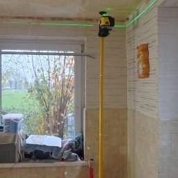 Usługi budowlane Vladysław Kliatskyi - Pomiar laserowy poziomu w trakcie prac remontowych w pomieszczeniu z oknem, widoczny laser na statywie, narzędzia i materiały budowlane.