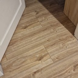 Usługi budowlane Vladysław Kliatskyi - Ułożone panele podłogowe z widoczną strukturą drewna, częściowo oświetlone, z fragmentem białej ościeżnicy i otwartych drzwi.