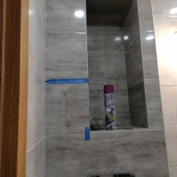 Usługi budowlane Vladysław Kliatskyi - Świeżo wykafelkowana ściana łazienki z wnęką, zabezpieczona niebieską taśmą malarską, widoczny fragment drewnianej ramy drzwi i dozownik mydła w tle.