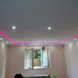 Wykończony sufit podwieszany z różowym oświetleniem LED wzdłuż krawędzi, punktowe lampy sufitowe, ściany pomalowane na jasny róż, widoczne okno z zielenią na zewnątrz, częściowo zakryte meble...