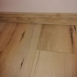 Jasne panele podłogowe imitujące drewno, z widocznymi słojami i detalami, ułożone przy listwie przypodłogowej w kolorze zbliżonym do podłogi i jasnej, jednolitej ścianie.