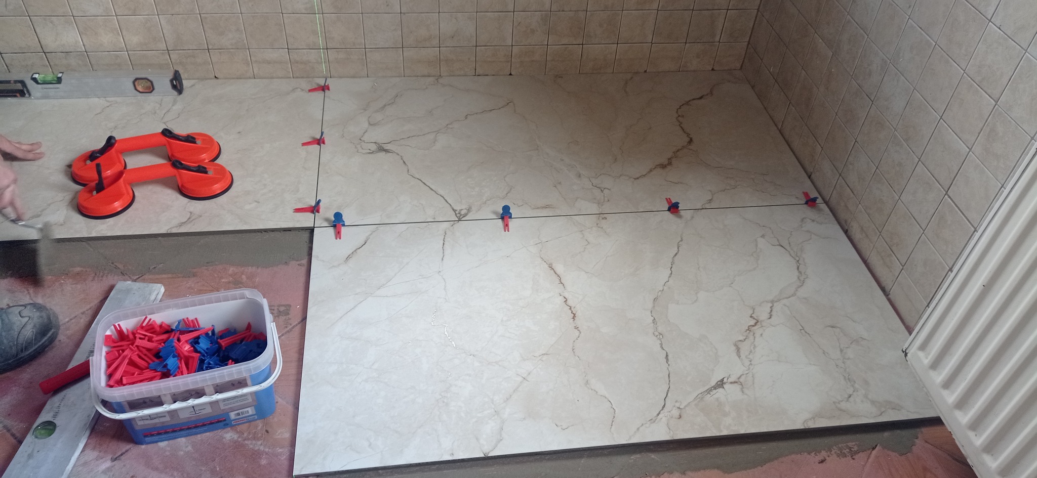 Proces układania beżowych płytek podłogowych imitujących marmur w łazience, z użyciem poziomicy, przyssawek do przenoszenia płytek i systemu poziomowania z czerwono-niebieskimi klipsami.