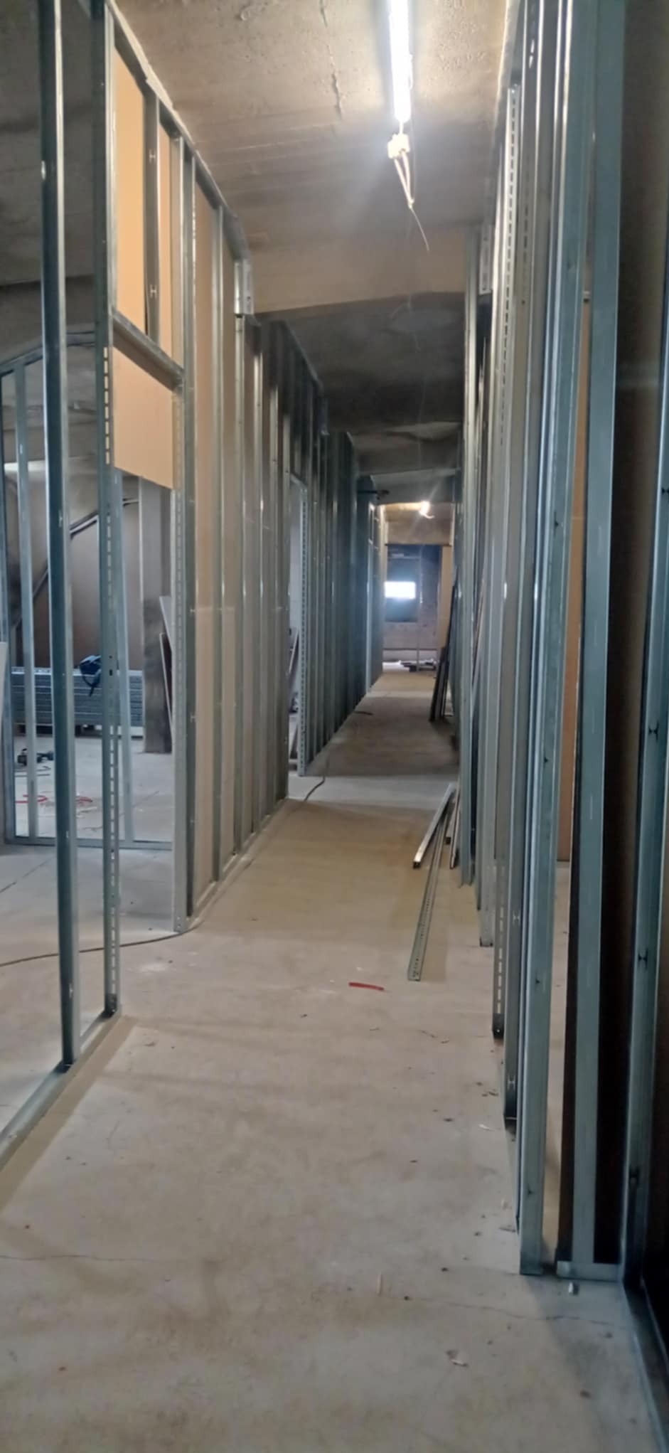 Korytarz w budynku w trakcie remontu, widoczne metalowe profile konstrukcyjne ścian działowych z częściowo zamontowanymi płytami. Świecąca lampa sufitowa w tle.