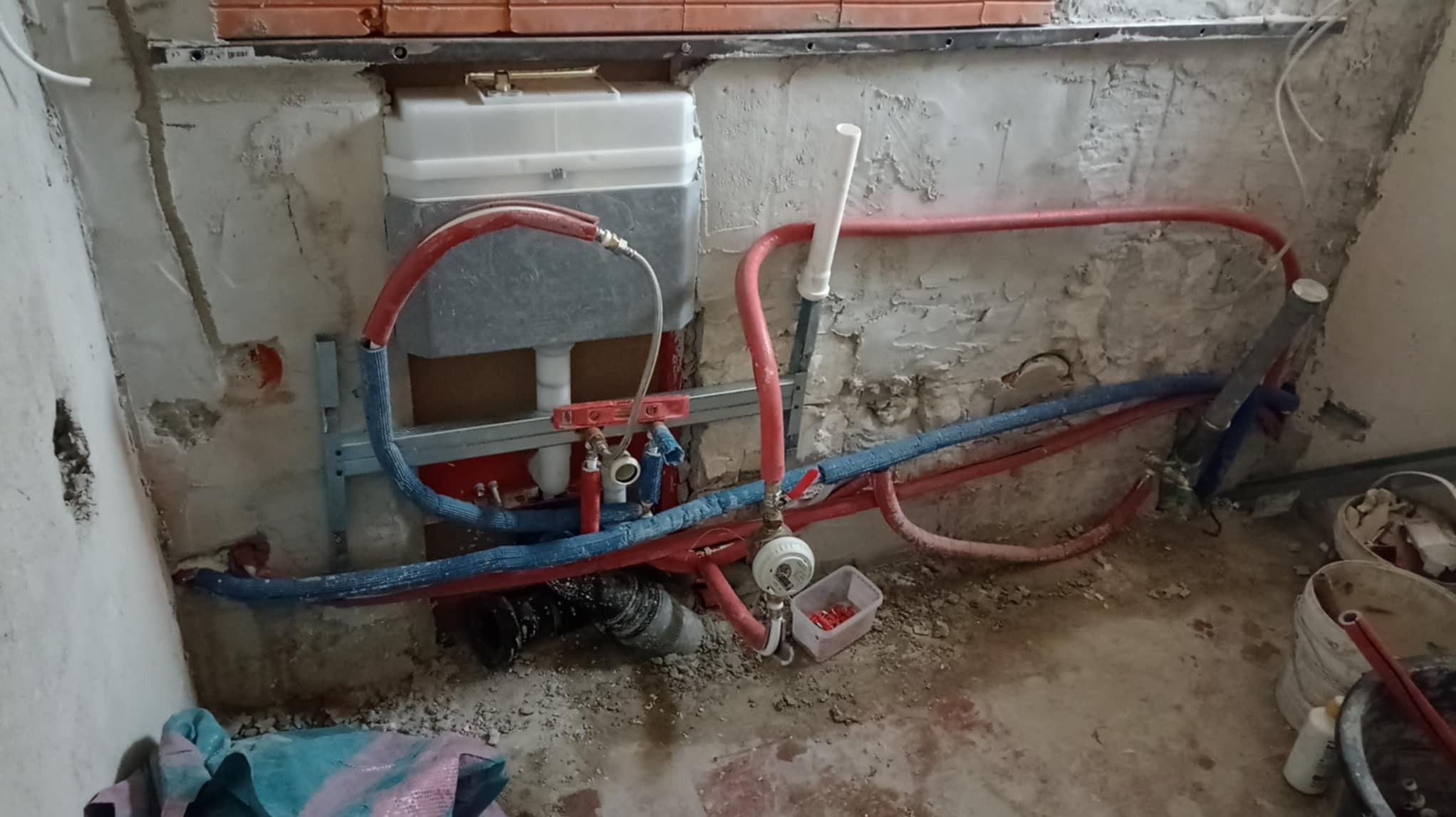 Stelaż podtynkowy WC zamontowany na surowej ścianie łazienki, widoczne rury doprowadzające wodę i odpływ, oraz drobne elementy instalacyjne w pudełku obok. Ściana wymaga dalszych prac wykończeniowych.