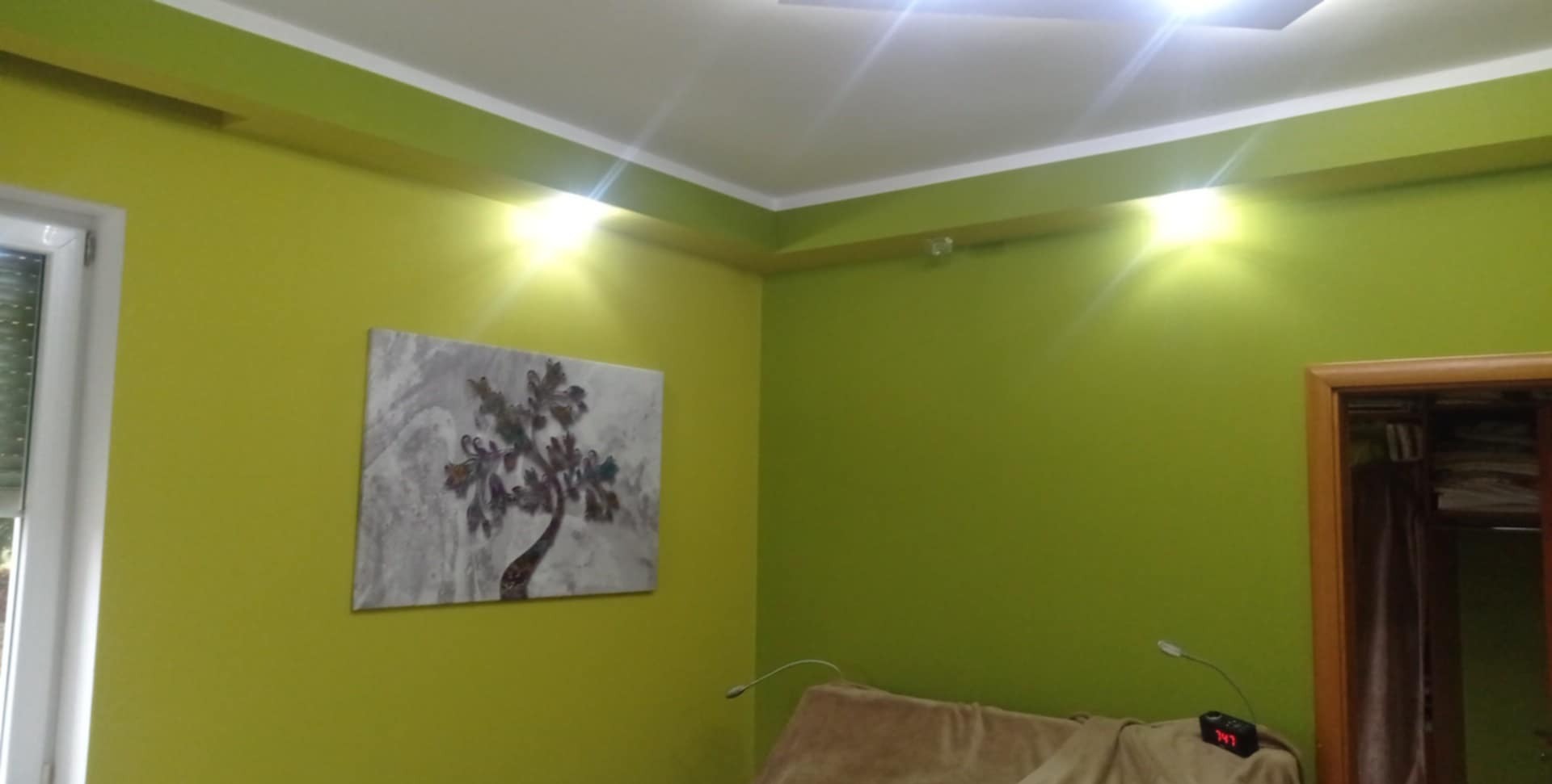 Ściana pomalowana na jasnozielony kolor z obrazem przedstawiającym drzewo, widoczny fragment sufitu z oświetleniem i fragment drzwi po prawej stronie.