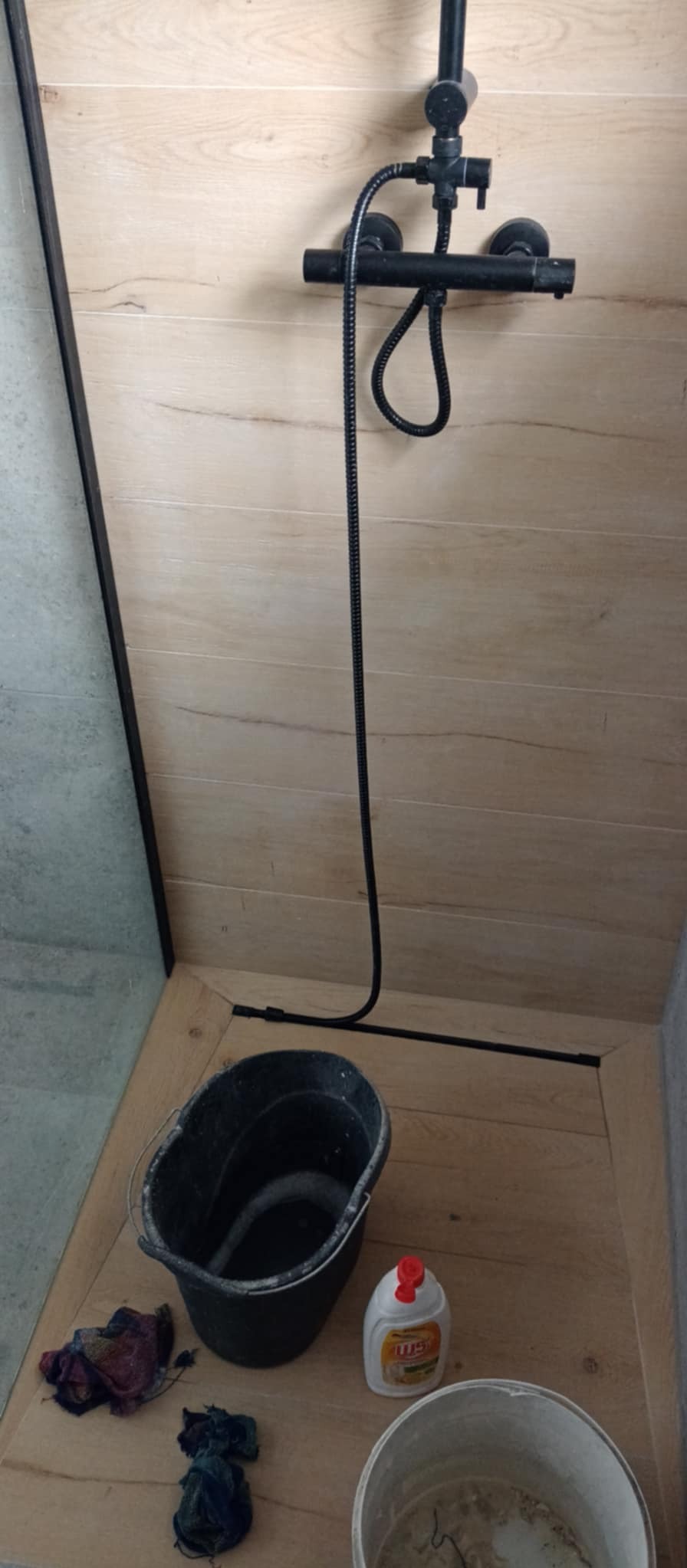 Fragment łazienki w trakcie remontu: widoczny czarny zestaw prysznicowy na tle drewnopodobnych płytek ściennych, na podłodze czarne wiadro z wodą, chemia czyszcząca i brudne ścierki.