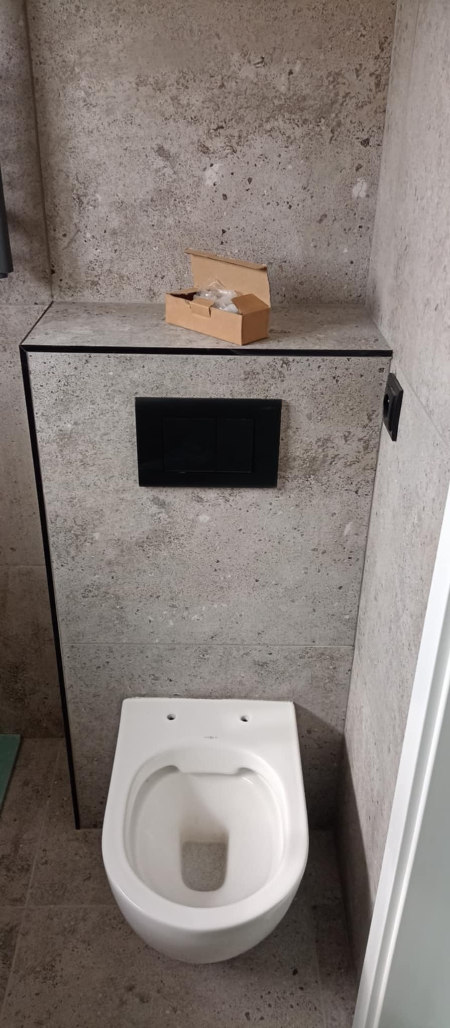 Świeżo zamontowany, podwieszany sedes z czarnym panelem spłukującym na tle ściany wykończonej płytami imitującymi beton, z otwartym pudełkiem z elementami montażowymi na górze