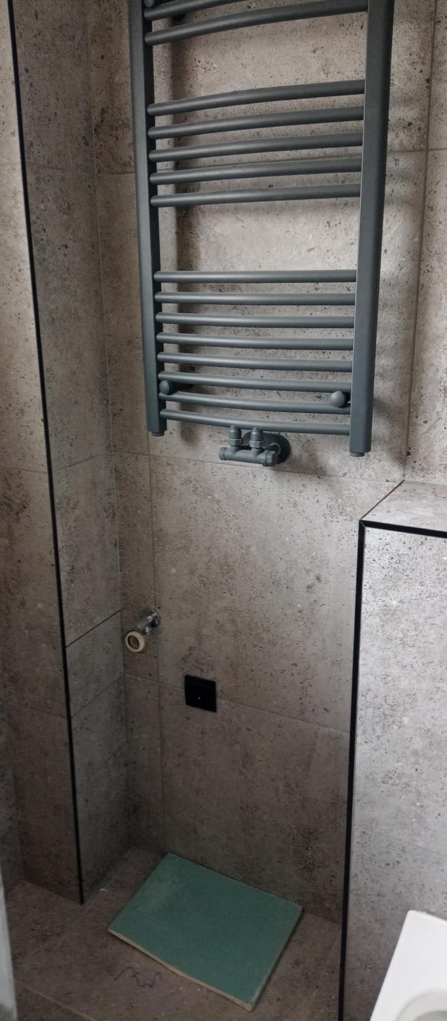 Szary grzejnik drabinkowy zamontowany na ścianie z płytek imitujących kamień w łazience, widoczny fragment podłogi z zieloną płytką, czarny włącznik i podejście wodne