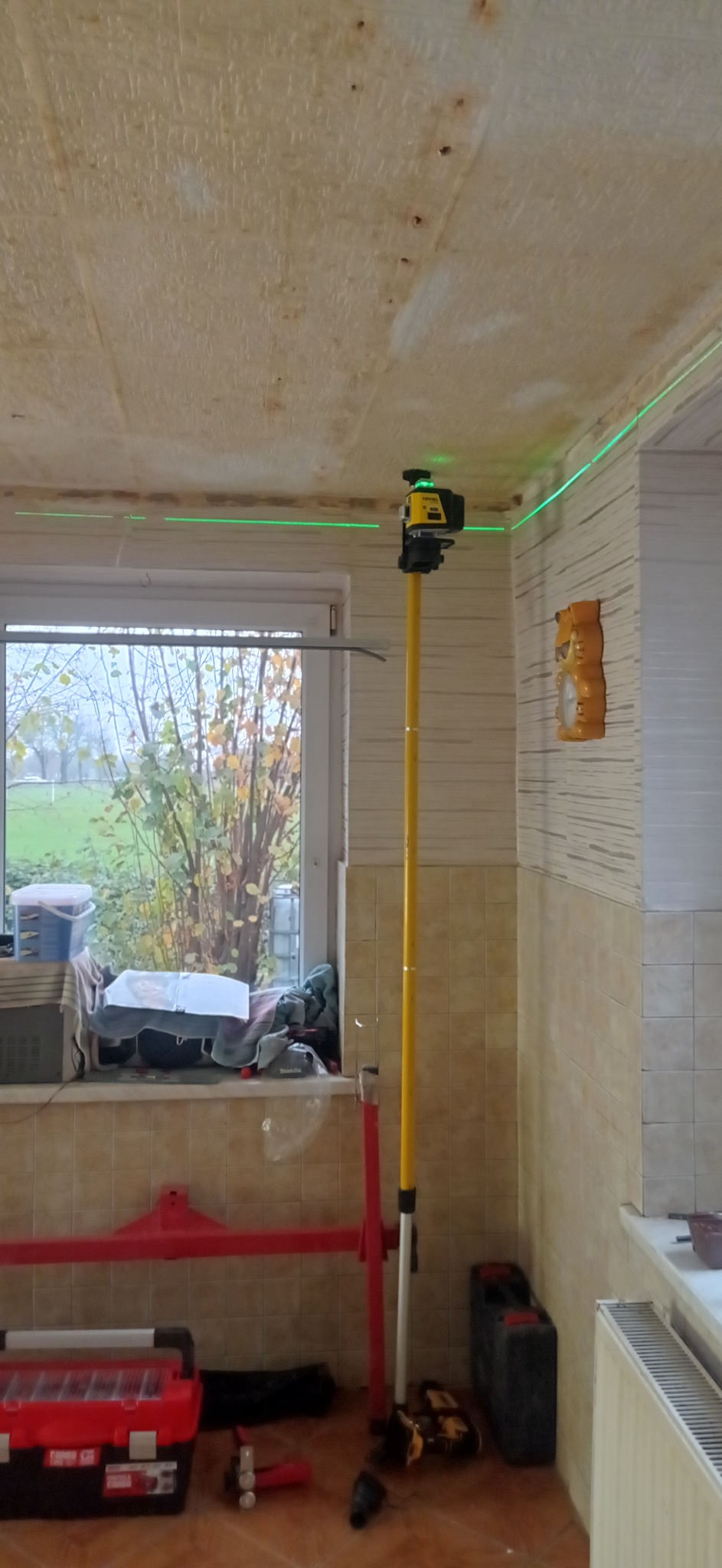 Pomiar laserowy poziomu w trakcie prac remontowych w pomieszczeniu z oknem, widoczny laser na statywie, narzędzia i materiały budowlane.