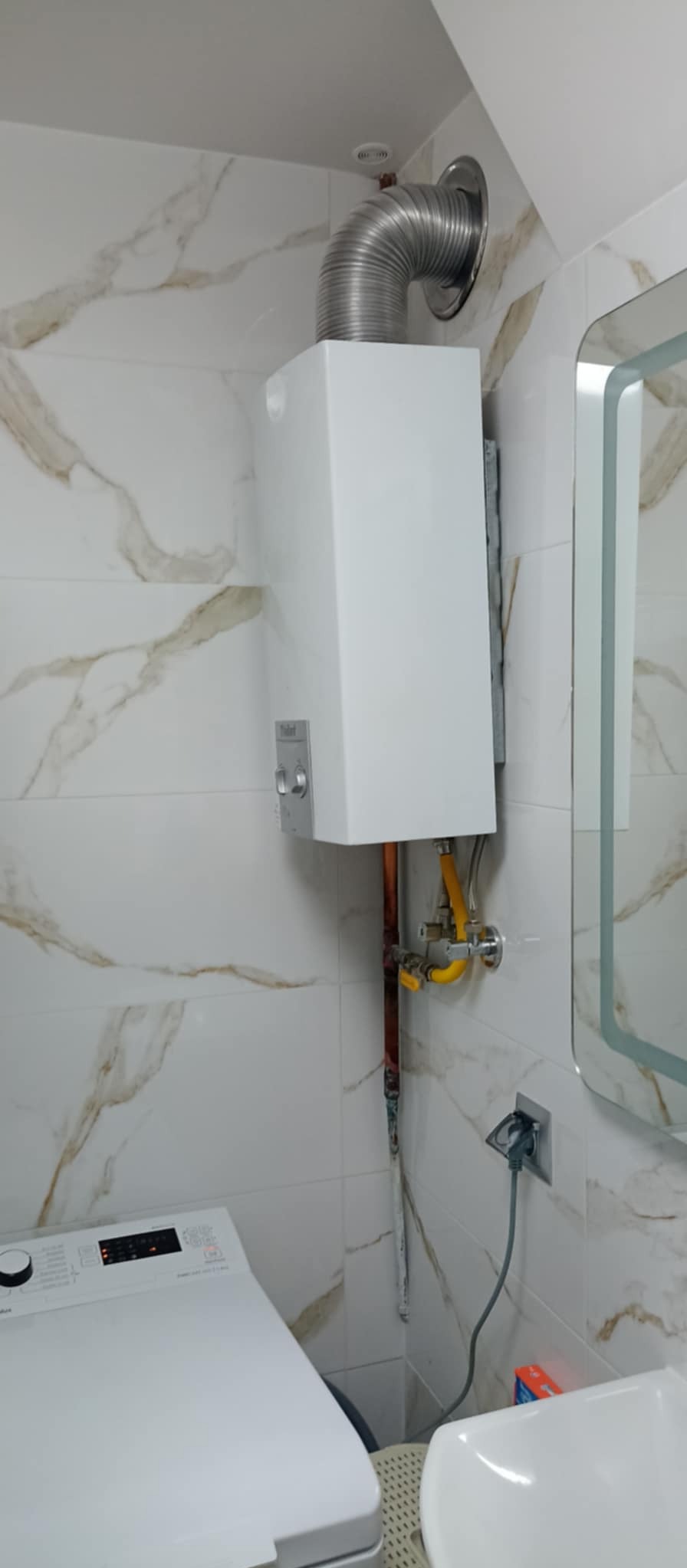 Biały piec gazowy z podłączonym kominem i instalacją, zamontowany na ścianie z marmurowymi płytkami w łazience, obok pralka i umywalka.