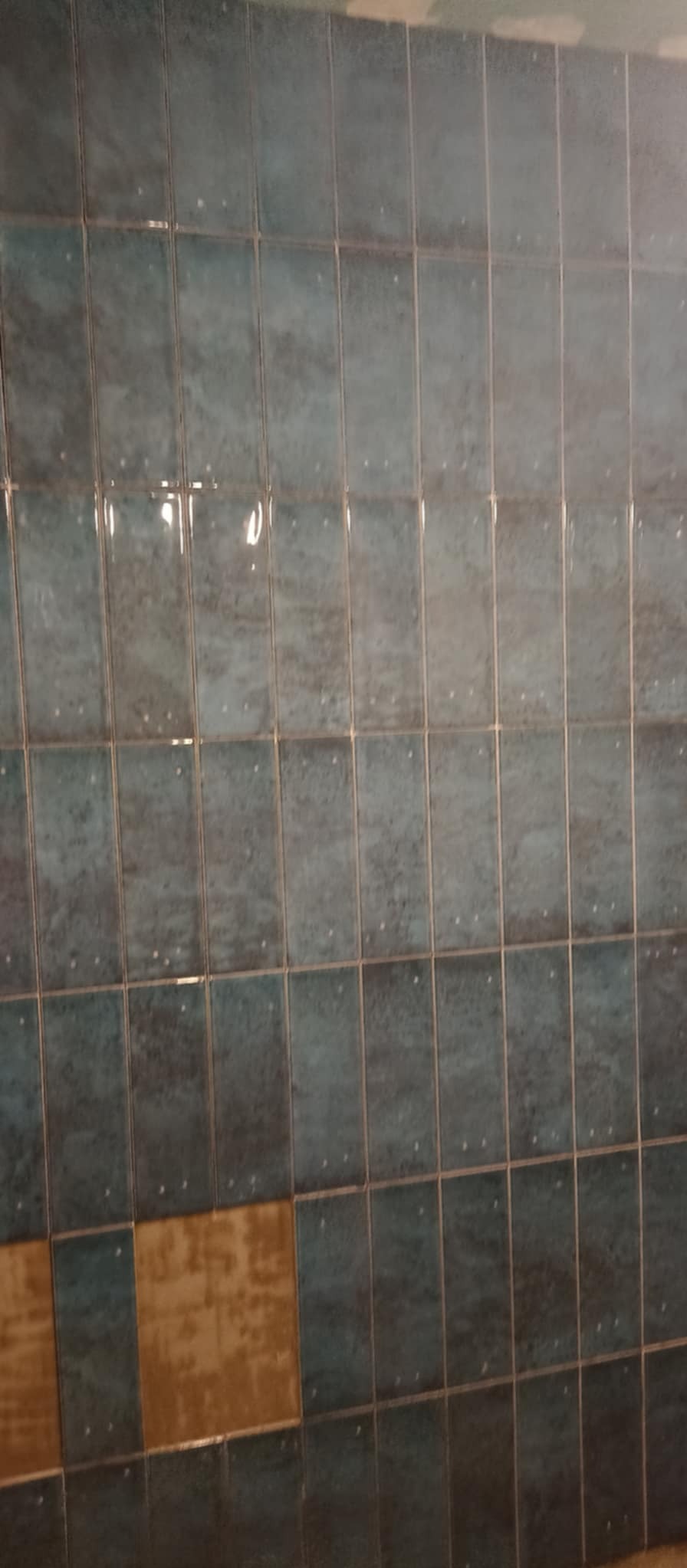 Fragment ściany z niebieskich, błyszczących płytek, z brakującą płytką w dolnym rzędzie, odsłaniającą żółtą warstwę pod spodem.