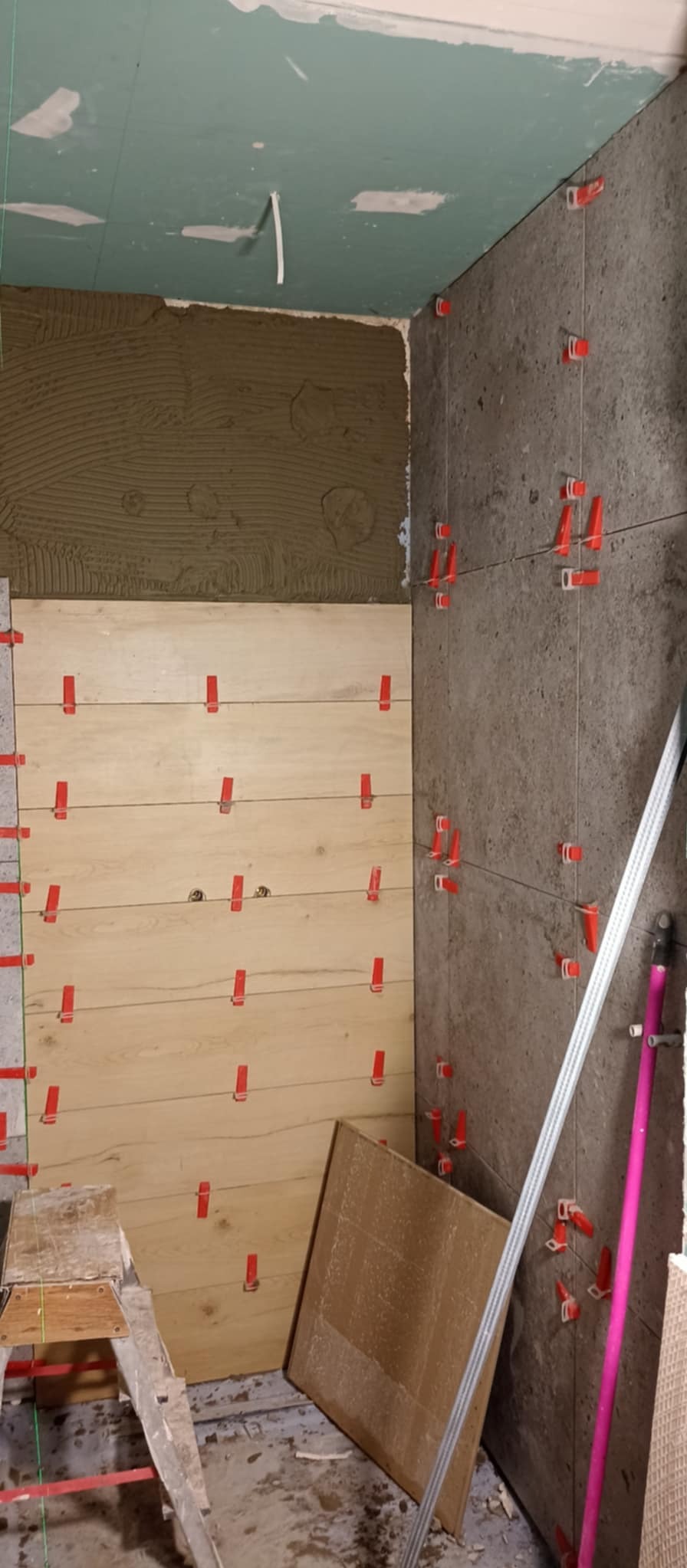 Ściana w trakcie wykańczania płytkami imitującymi drewno i kamień, z widocznymi krzyżykami dystansowymi, klejem na ścianie i poziomnicą opartą o ścianę, ukazująca etap prac w pomieszczeniu.