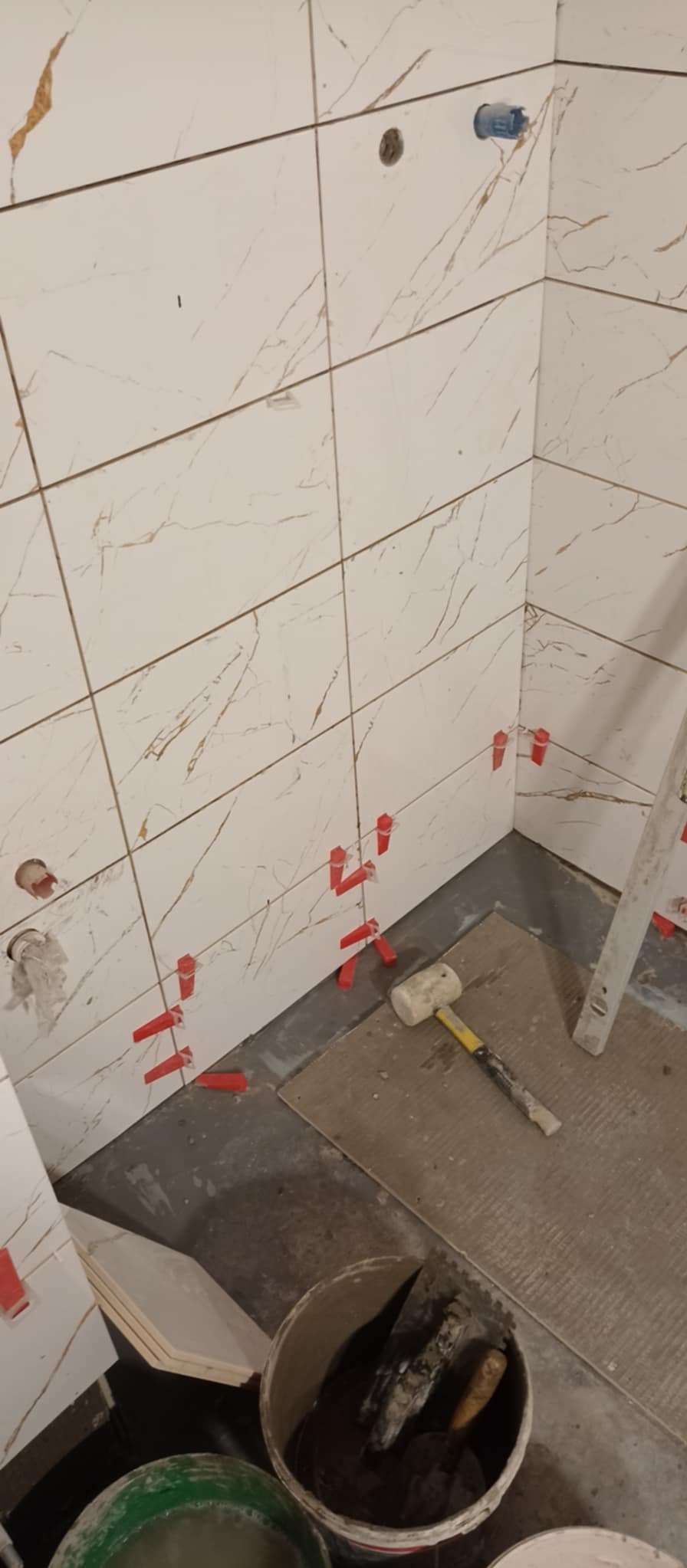 Fragment ściany łazienki w trakcie układania płytek imitujących marmur z użyciem czerwonych klipsów poziomujących, widoczny gumowy młotek, poziomica i wiadro z zaprawą.