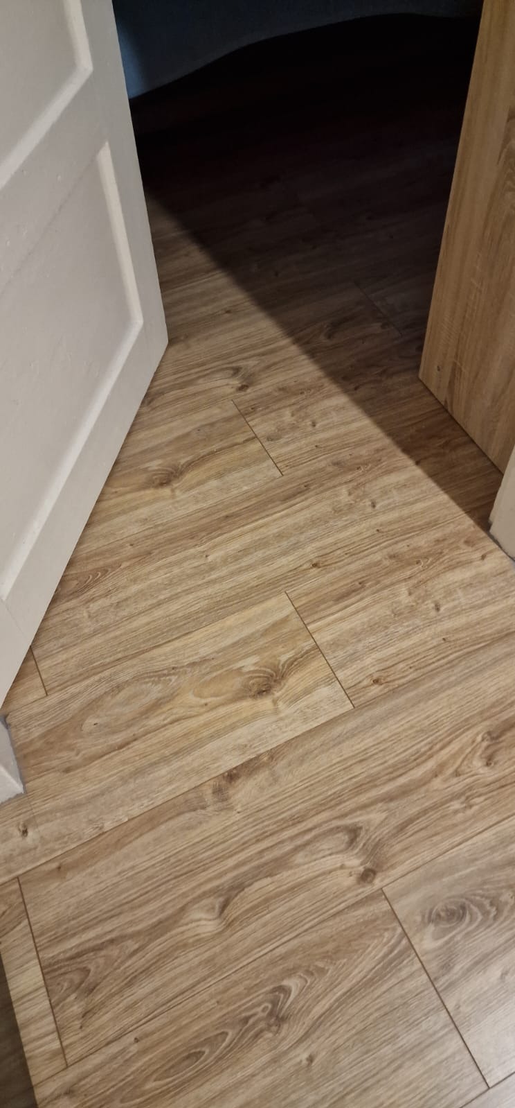 Ułożone panele podłogowe z widoczną strukturą drewna, częściowo oświetlone, z fragmentem białej ościeżnicy i otwartych drzwi.