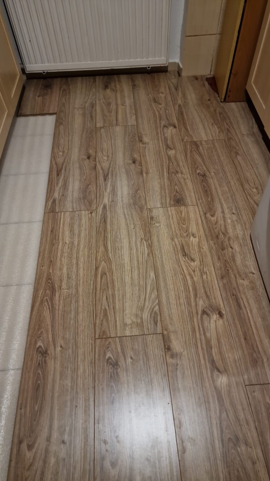 Ułożone panele podłogowe o wzorze drewna, widoczne fragmenty białej podłogi i grzejnika.