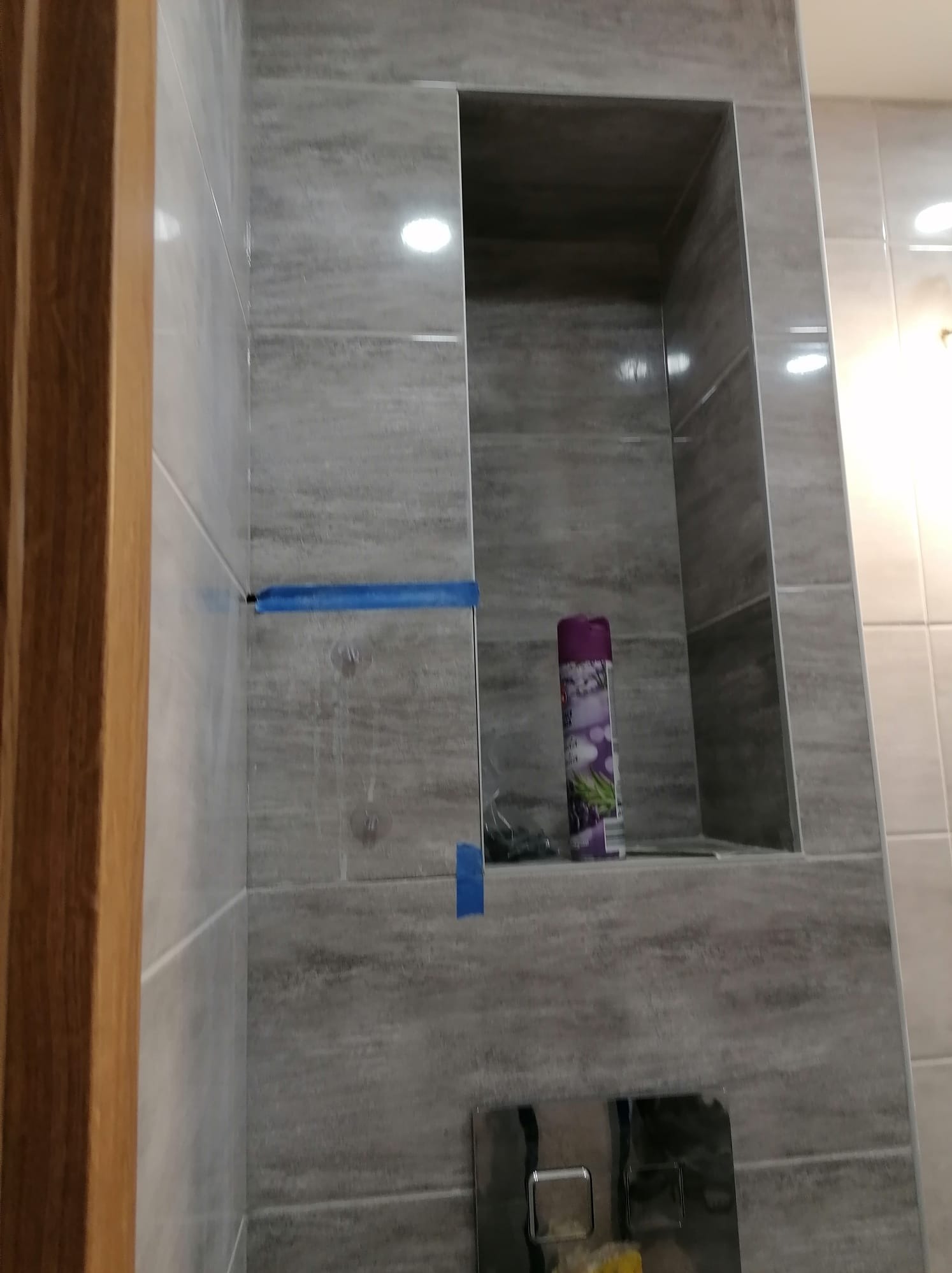 Świeżo wykafelkowana ściana łazienki z wnęką, zabezpieczona niebieską taśmą malarską, widoczny fragment drewnianej ramy drzwi i dozownik mydła w tle.