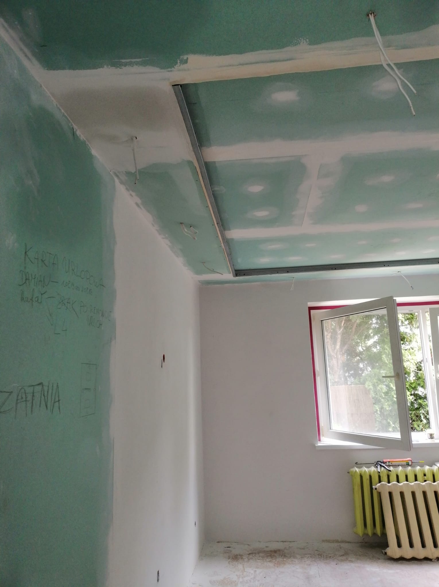 Wnętrze w trakcie remontu: ściana pokryta zieloną płytą, z napisem 'Karta urlopowa', sufit z zamontowaną konstrukcją metalową, otwarte okno z widokiem na zieleń, stary grzejnik przy ścianie.