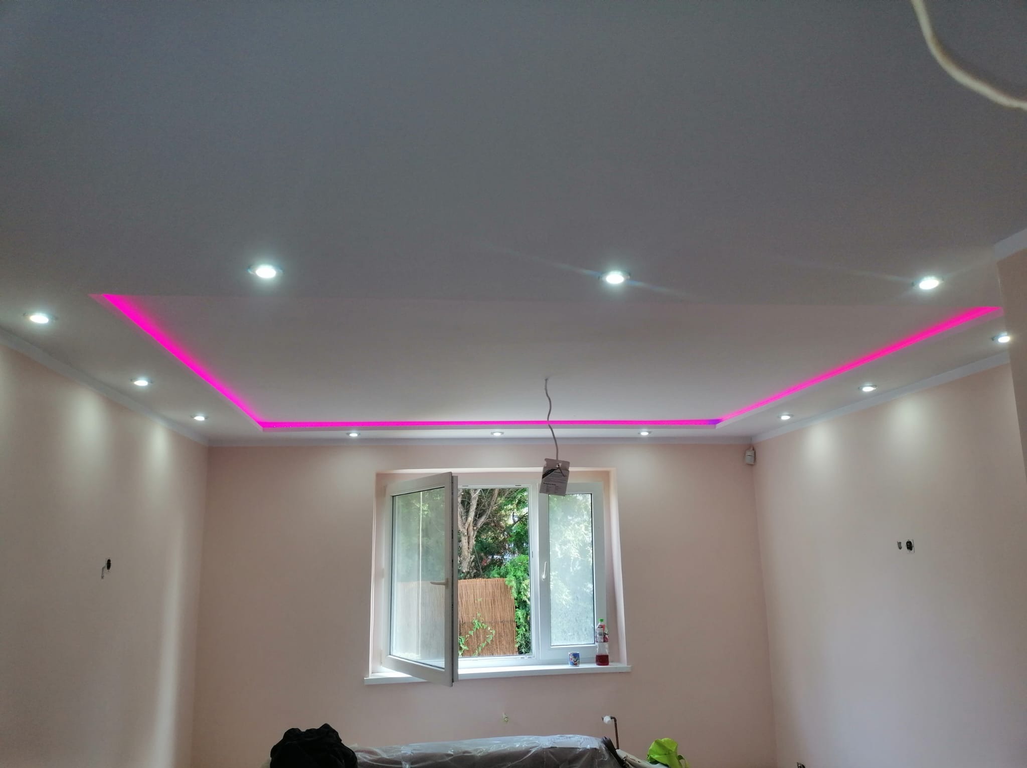 Wykończony sufit podwieszany z różowym oświetleniem LED wzdłuż krawędzi, punktowe lampy sufitowe, ściany pomalowane na jasny róż, widoczne okno z zielenią na zewnątrz, częściowo zakryte meble...