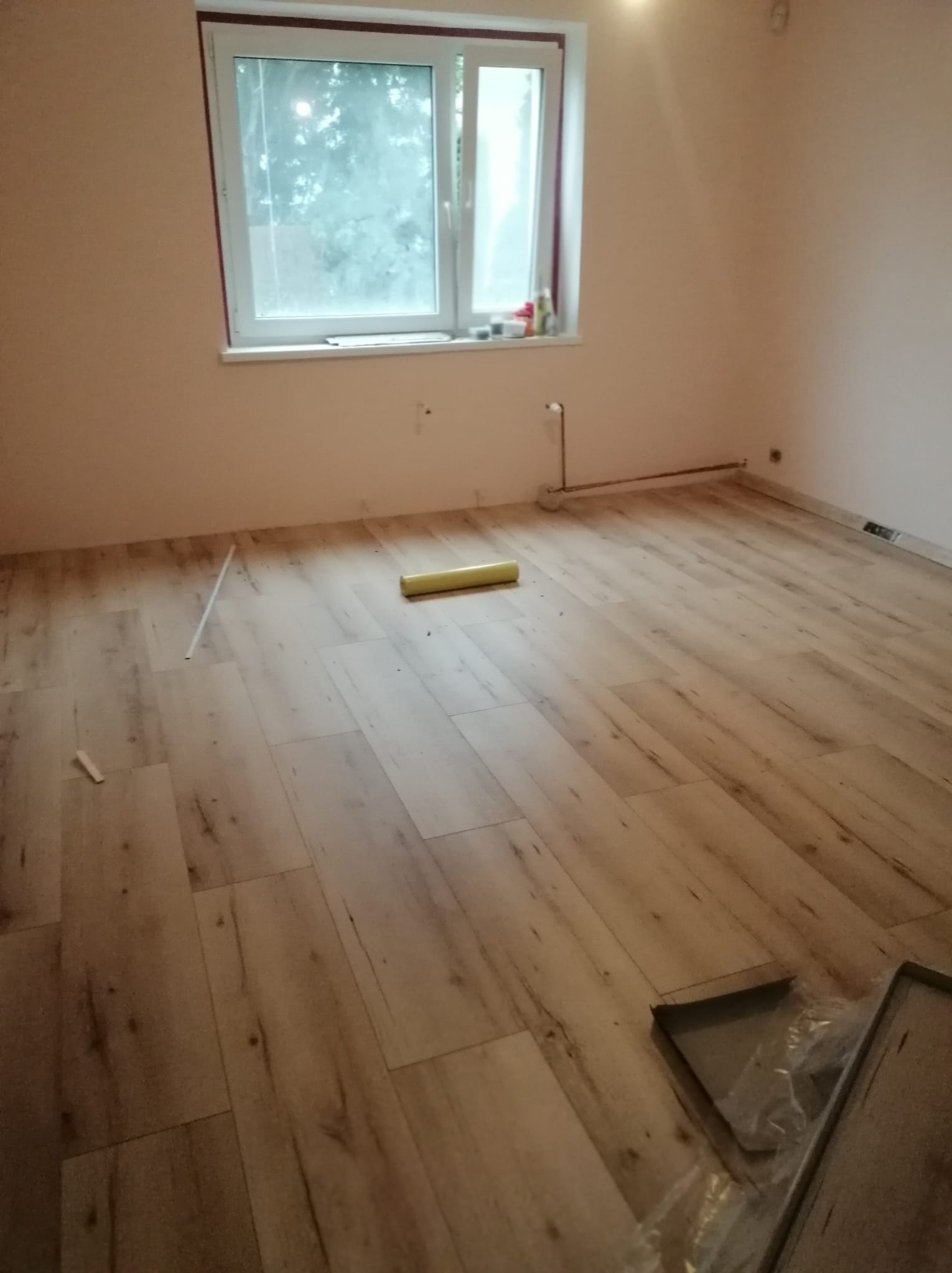 Jasne panele podłogowe z widoczną fakturą drewna, częściowo ułożone w pokoju z oknem, żółty rulon podkładu i narzędzia w rogu.