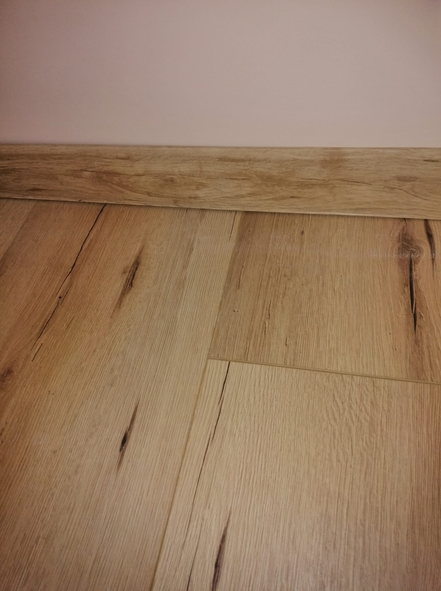 Jasne panele podłogowe imitujące drewno, z widocznymi słojami i detalami, ułożone przy listwie przypodłogowej w kolorze zbliżonym do podłogi i jasnej, jednolitej ścianie.