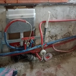 Usługi budowlane Wladysław Kliatskyi - Montaż podtynkowego stelaża WC na surowej ścianie, widoczne rury instalacyjne w kolorze czerwonym i niebieskim, oraz biała spłuczka. Ściana wymaga dalszych prac wykończeniowych.