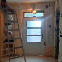 Usługi budowlane Wladysław Kliatskyi - Mężczyzna na drewnianej drabinie podczas prac przygotowawczych do malowania ścian w pomieszczeniu z widoczną starą instalacją elektryczną i fragmentami usuniętej farby na ścianach.