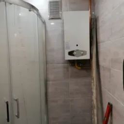 Nowoczesna łazienka w trakcie wykańczania: zamontowany piec gazowy, widoczna kabina prysznicowa, szare płytki, pionowa poziomica oparta o ścianę.