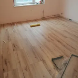 Układanie paneli podłogowych w jasnym pokoju z oknem, widoczne narzędzia i rolka folii izolacyjnej.