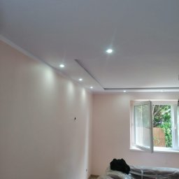 Usługi budowlane Wladysław Kliatskyi - Jasne wnętrze po remoncie: gładkie, pomalowane ściany w odcieniach różu, podwieszany sufit z oświetleniem punktowym, okno z widokiem na zieleń, meble zabezpieczone folią ochronną.