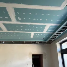 Niebieski sufit z płyt kartonowo-gipsowych, częściowo zaszpachlowany, z widoczną metalową konstrukcją nośną i instalacją elektryczną. W tle surowa ściana i fragment okna.