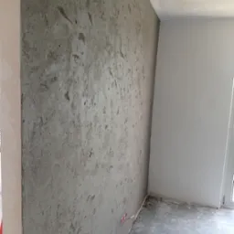 Surowa ściana z efektem betonu w rogu pokoju, widoczne niedoskonałości i faktura, surowy beton na podłodze, przygotowanie do dalszych prac wykończeniowych.
