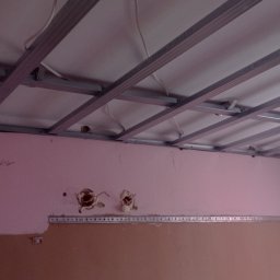 Usługi budowlane Wladysław Kliatskyi - Montaż metalowej konstrukcji sufitu podwieszanego z płyt gipsowo-kartonowych w pomieszczeniu z różowymi ścianami i widocznymi instalacjami elektrycznymi.