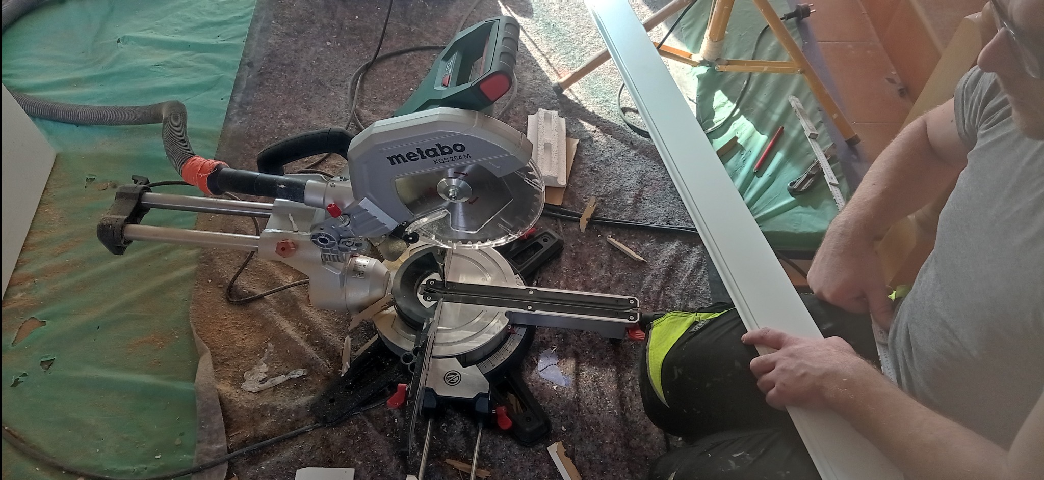 Pracownik przycina białą listwę przypodłogową za pomocą piły ukośnicy Metabo KGS 254 M na tle folii ochronnej, widoczne drobne odpady i narzędzia.