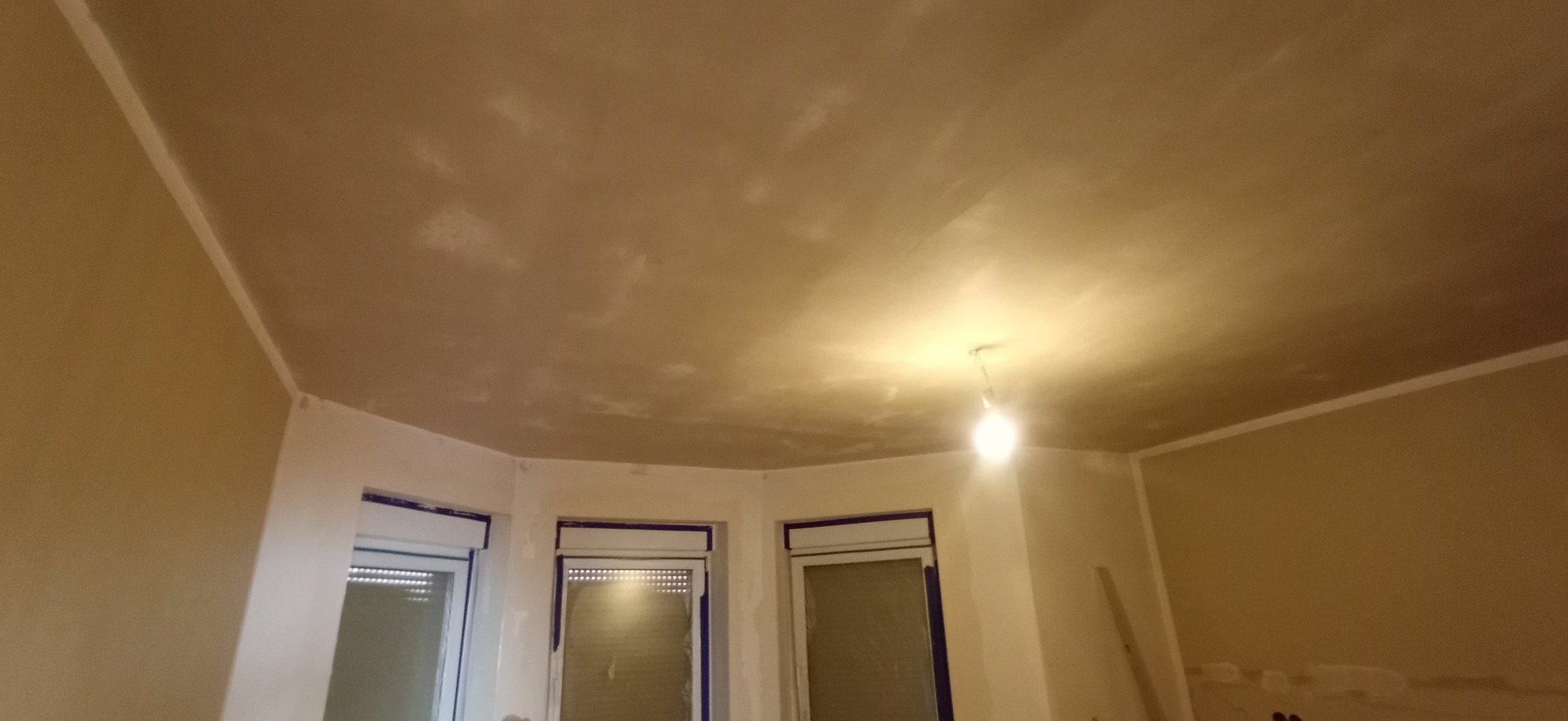 Niedokończona powierzchnia sufitu po nałożeniu szpachli, widoczne nierówności i miejsca wymagające dalszej obróbki, z żarówką na kablu jako oświetlenie tymczasowe.