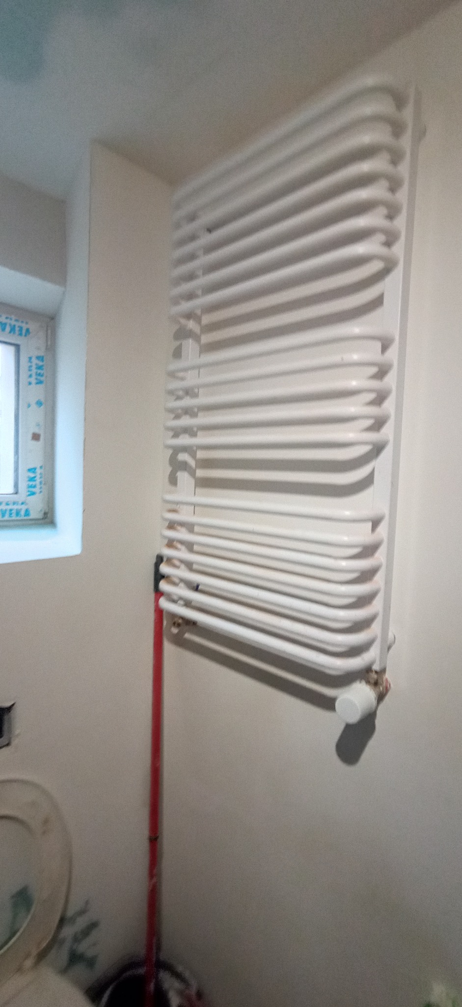 Biały, drabinkowy grzejnik łazienkowy zamontowany na ścianie, widoczne rury doprowadzające wodę, okno z ramą PCV w tle, fragment muszli klozetowej w dolnym lewym rogu.