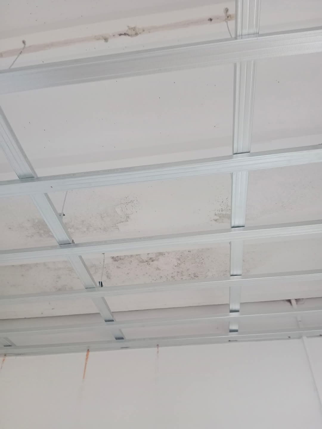 Stelaż sufitowy z metalowych profili, widoczne plamy wilgoci na oryginalnym suficie, przygotowanie do montażu płyt.