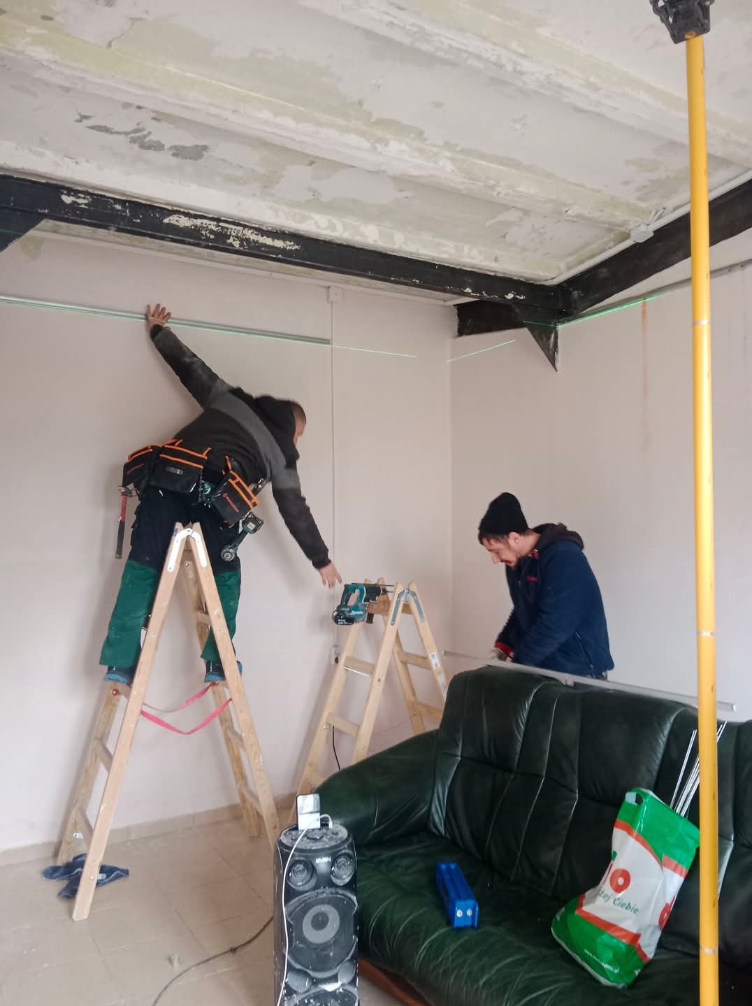 Dwóch mężczyzn pracuje w pomieszczeniu, jeden na drabinie wyznacza poziom laserem na ścianie, drugi przycina listwę, widoczny stary sufit, narzędzia i głośnik na podłodze.