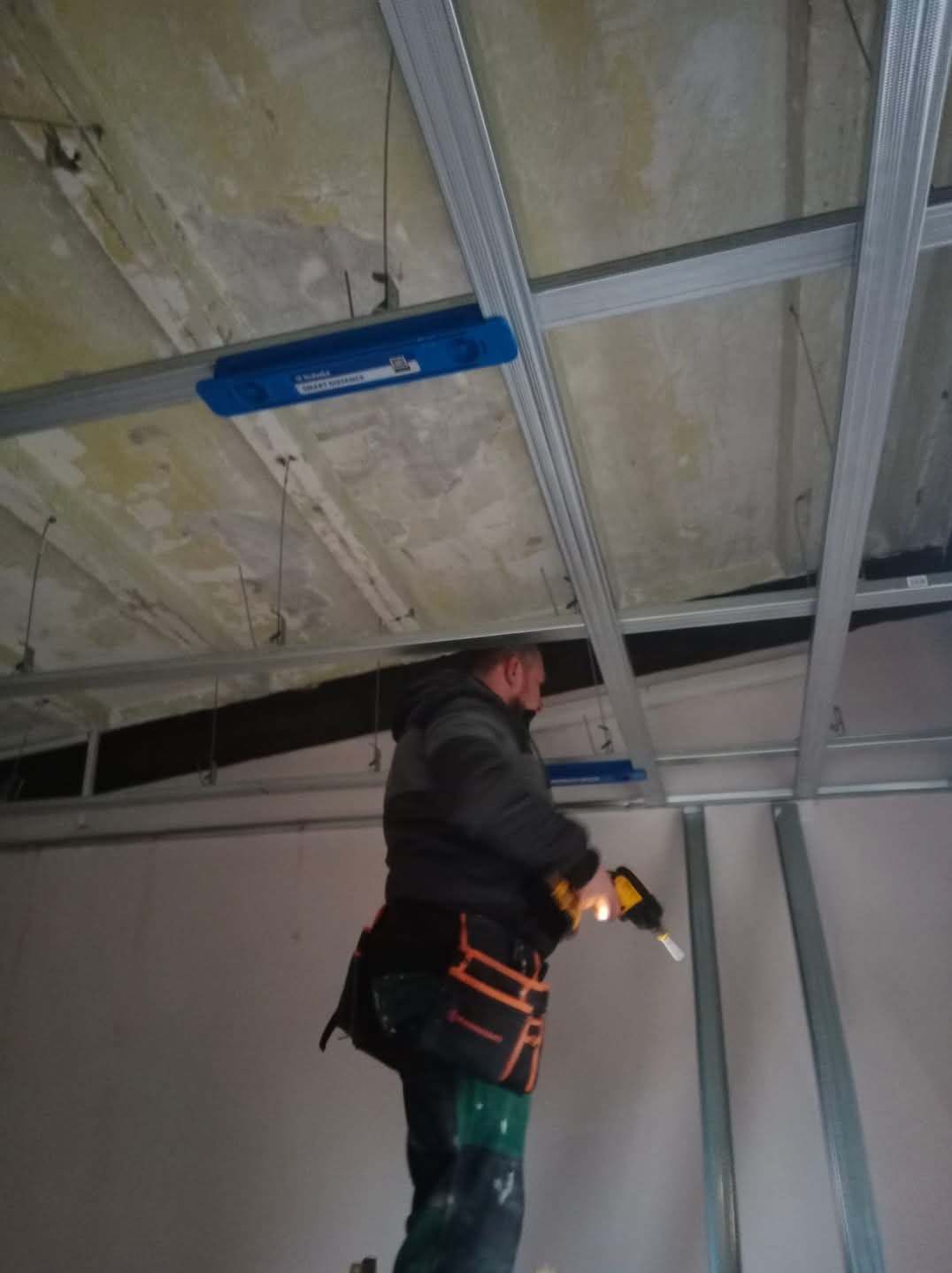 Montaż metalowego stelaża sufitu podwieszanego z płyty karton gips, widoczne profile, uchwyty, pracownik z wkrętarką w trakcie pracy.