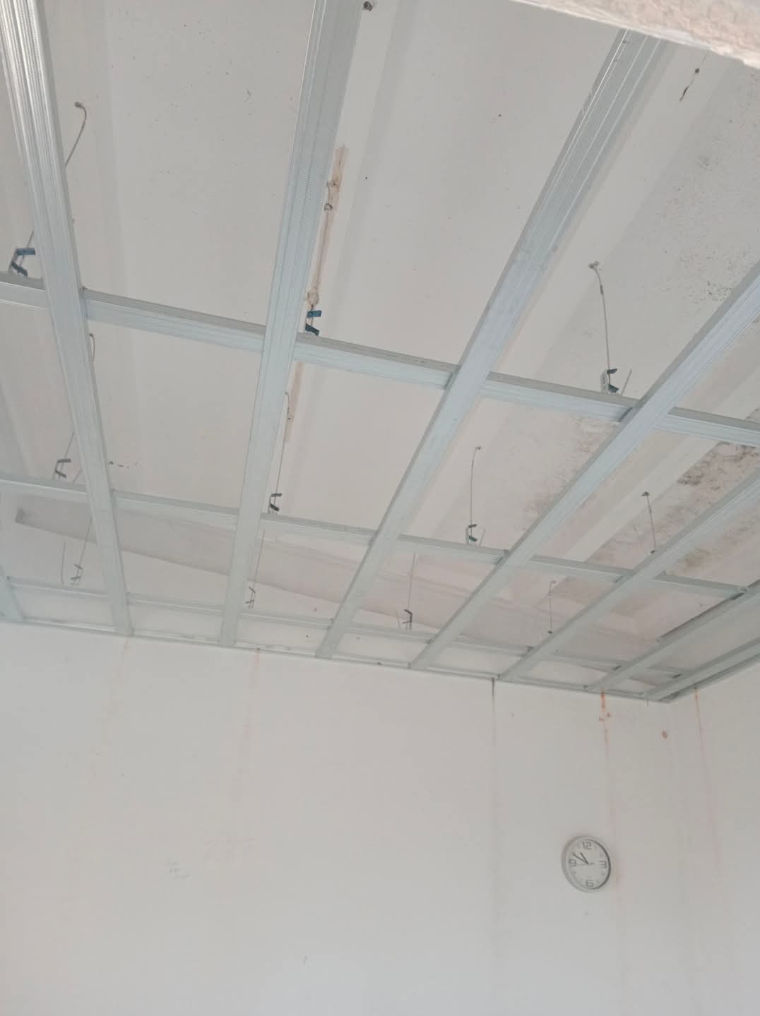 Metalowy stelaż sufitowy z widocznymi elementami mocującymi, przygotowany do montażu płyt, na ścianie widoczny zegar.