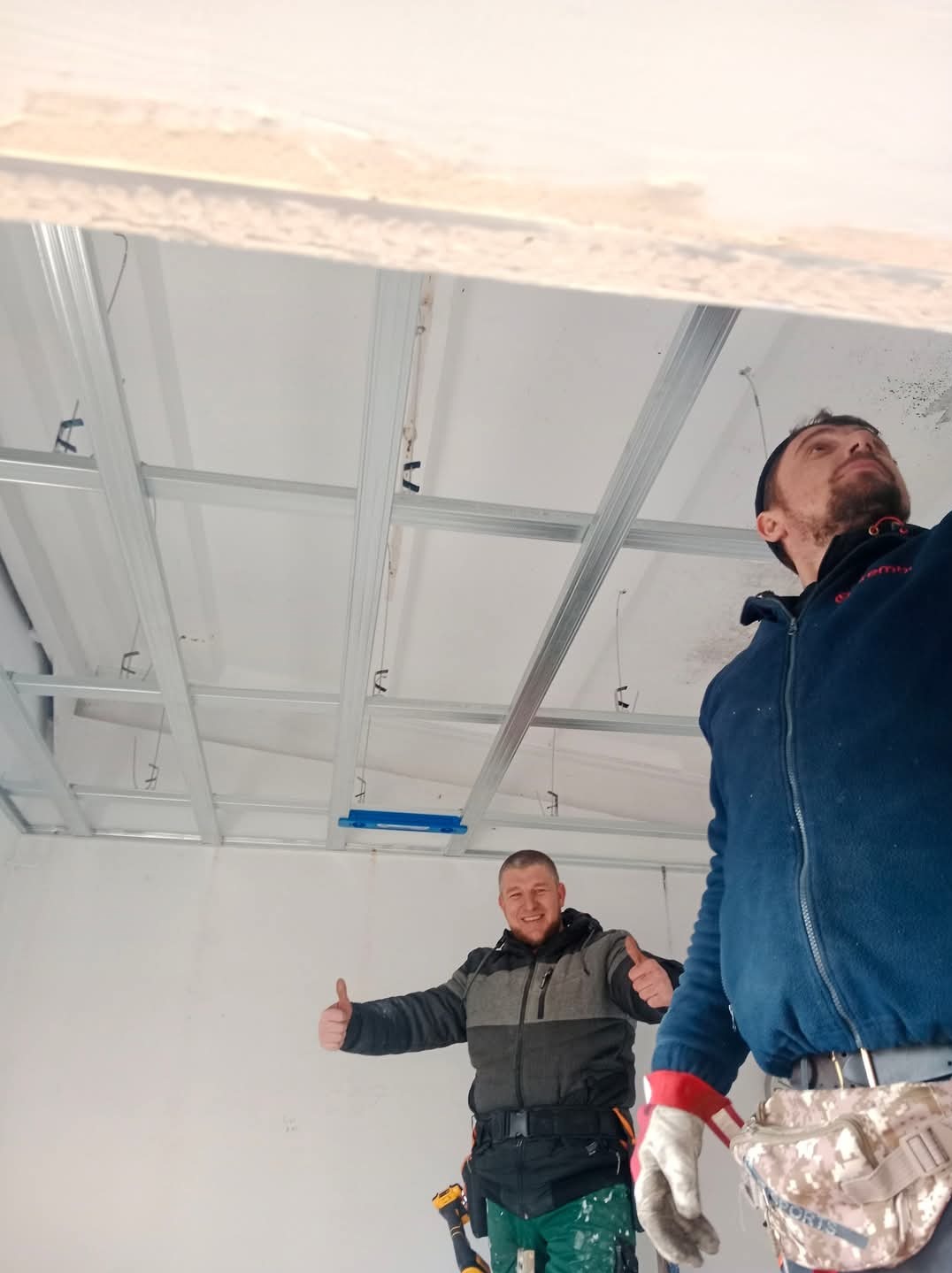 Dwaj uśmiechnięci monterzy prezentują metalową konstrukcję sufitu podwieszanego, jeden z nich z uniesionymi kciukami, widoczne narzędzia przy pasie.