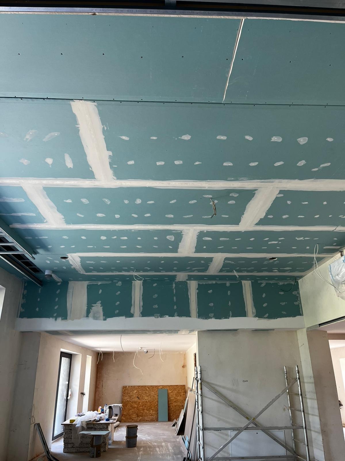 Niebieskie płyty sufitowe, częściowo zaszpachlowane, z widocznymi otworami i przewodami. W tle widoczne rusztowanie i materiały budowlane.