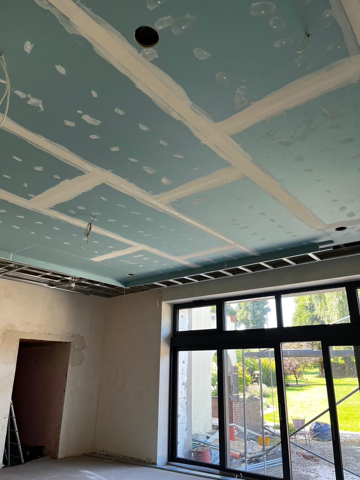 Niebieski sufit z płyt gipsowo-kartonowych w trakcie prac wykończeniowych, widoczne spoiny i otwory na oświetlenie, metalowa konstrukcja podwieszana, okno z widokiem na zieleń.