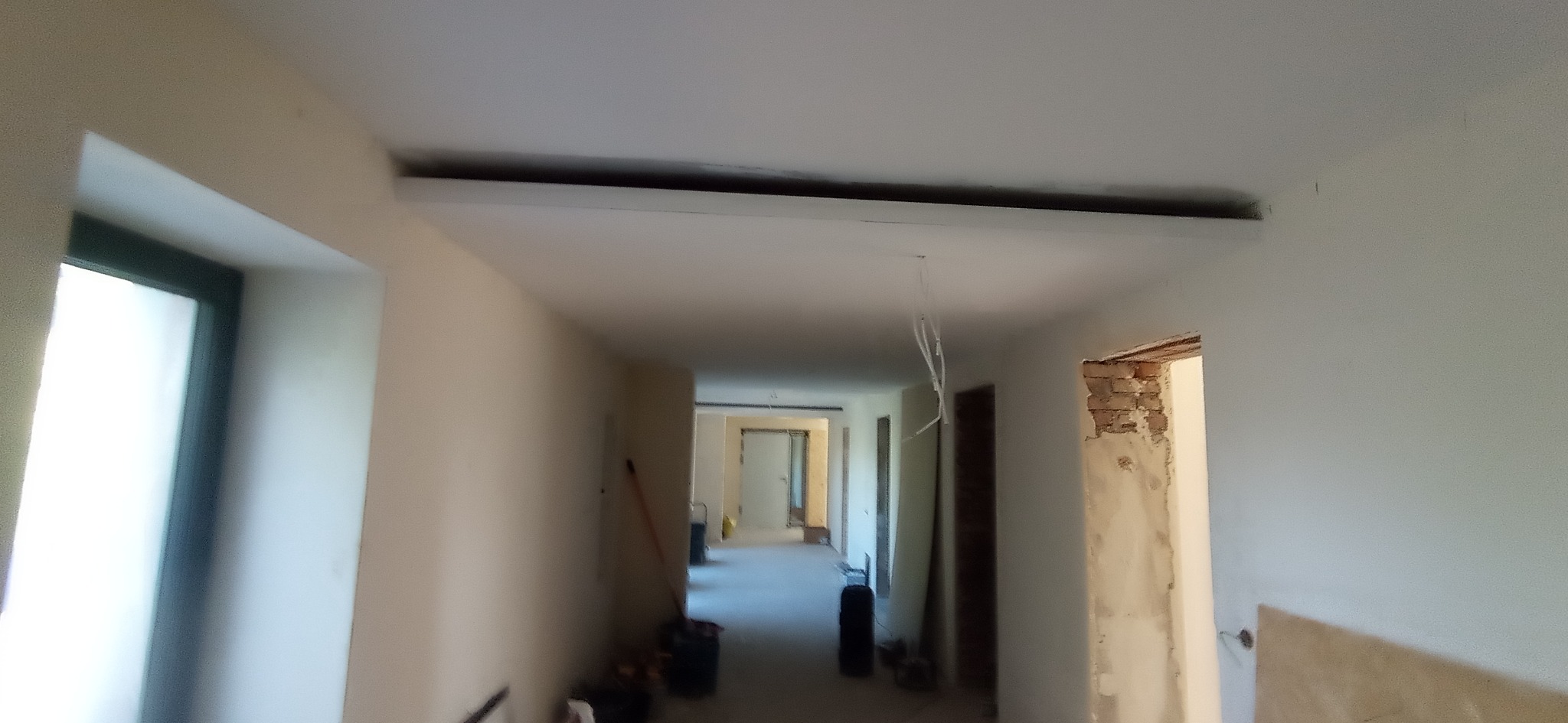 Korytarz w trakcie remontu: świeżo pomalowane ściany na jasny odcień, widoczne przygotowania do montażu oświetlenia sufitowego i odsłonięta cegła wokół ościeżnicy.
