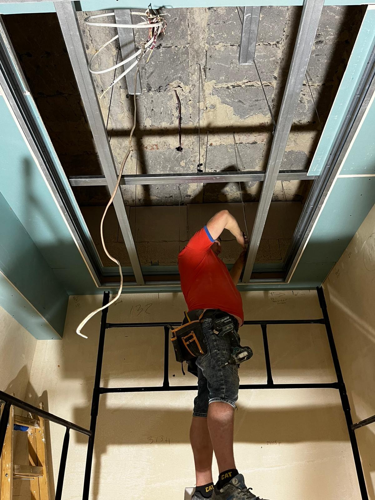 Monterażysta w czerwonej koszulce mocuje metalowy stelaż sufitu podwieszanego z płyt g-k, widoczne przewody i fragment ściany z zamontowaną płytą.