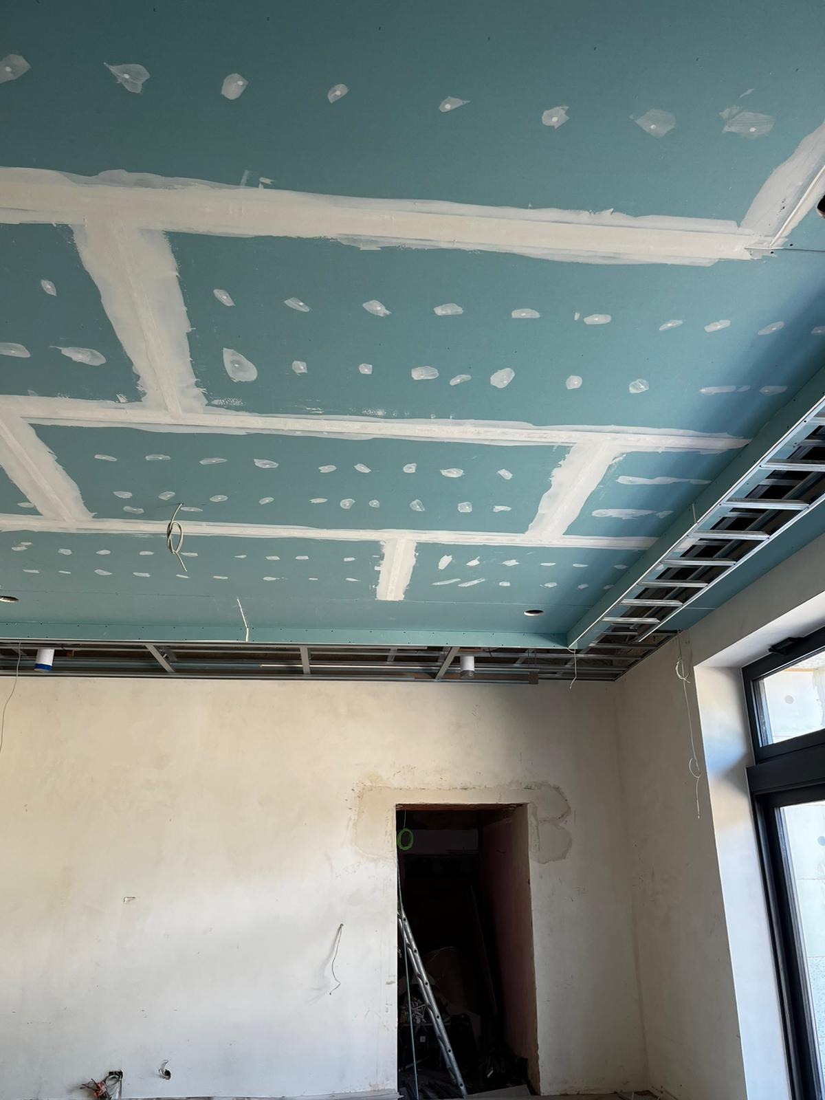 Niebieski sufit z płyt kartonowo-gipsowych, częściowo wyszpachlowany, z widoczną metalową konstrukcją nośną i przewodami. Ściany w trakcie przygotowania do dalszych prac.