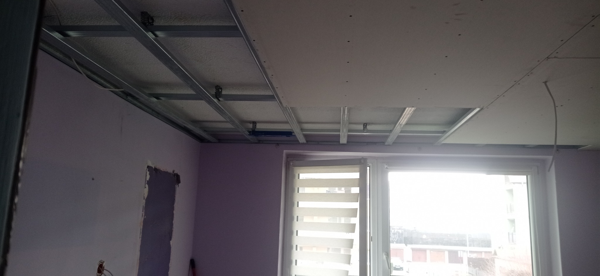 Montaż sufitu podwieszanego z płyt kartonowo-gipsowych na metalowej konstrukcji w pokoju z oknem. Widoczne profile i częściowo zamontowane płyty.